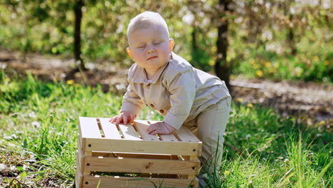 lovely serious blond baby boy stands leaning on the wooden crate. cute caucasian child outdoors in spring.