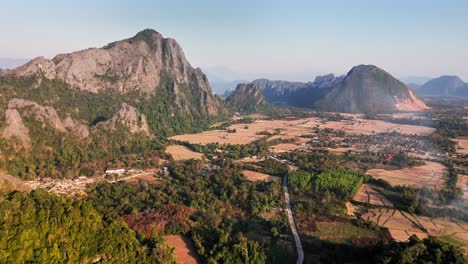 expansive drone shot capturing vang vieng’s dramatic karst mountains, rural farmlands, and winding roads in northern laos during late afternoon with warm golden tones