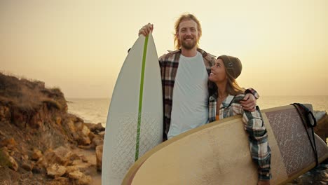 Portrait-of-a-happy-couple,-a-blond-guy-with-a-beard-in-a-checkered-shirt-stands-and-hugs-his-blonde-girlfriend-in-a-green-hat.-A-guy-and-a-girl-hold-surfboards-and-stand-on-a-rocky-seashore-in-the-morning