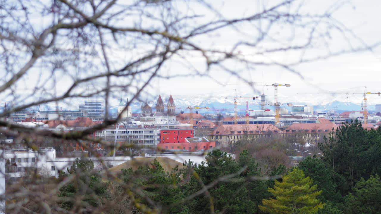 A panoramic view of Munich's skyline with historic architecture and construction cranes, framed by tree branches. Snow-capped Alps in the background under a cloudy winter sky, showcasing urban