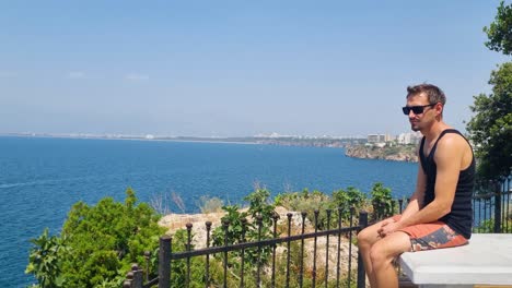 dressed in summer clothes and sunglasses, a man relaxes on the cliffs of antalya’s coast, taking in the sweeping, wind-filled view of the mediterranean on a bright spring day