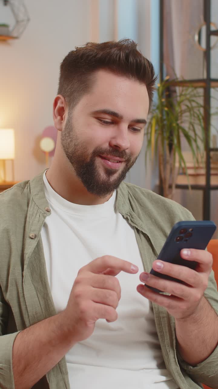 Happy young man sitting on sofa using smartphone texting smiling at home living room apartment