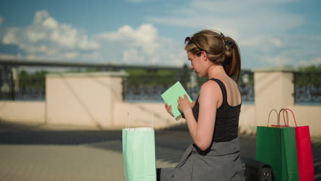 woman seated on outdoor with shopping bags on both sides, she retrieves a book from mint green bag, opens it, and flips through the pages, blurred background shows fence and distant bridge