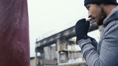 side view of caucasian man in black beanie hitting a punching bag outdoors an abandoned factory on a cloudy morning