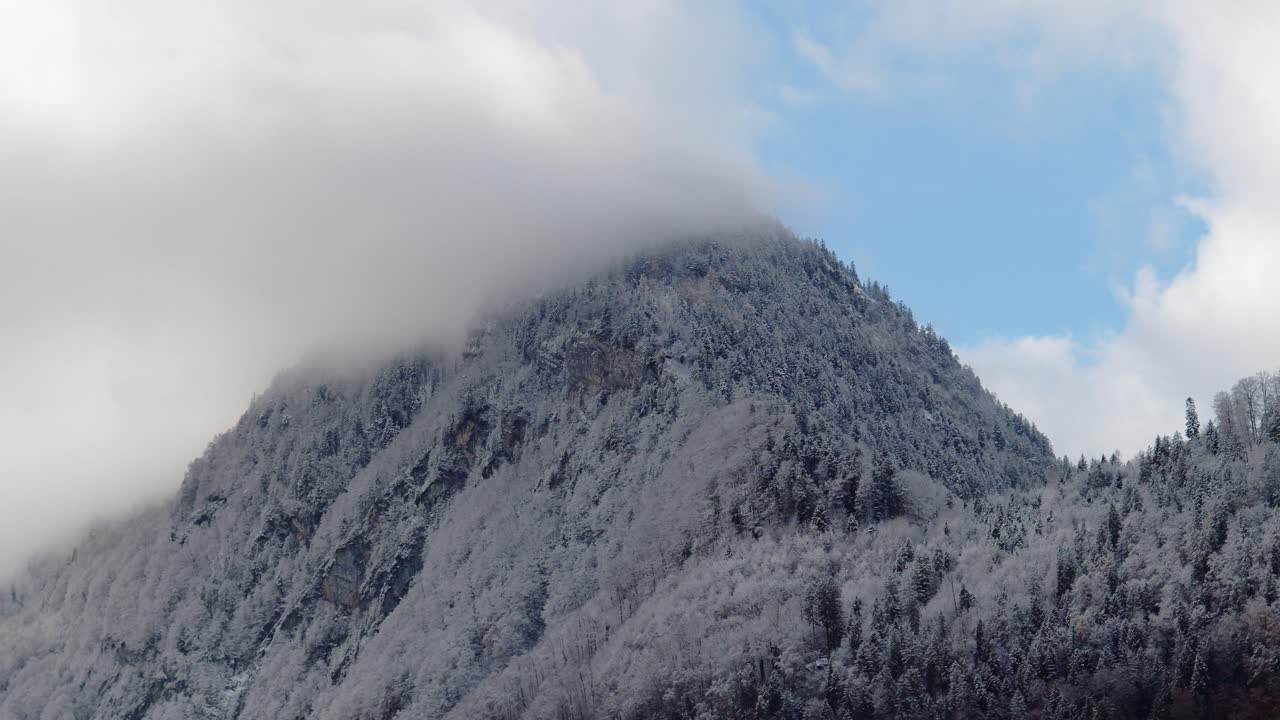 Snow-covered mountain with its top wrapped in fog under a partially blue sky. The scene captures a calm alpine winter atmosphere with clouds meeting rugged slopes