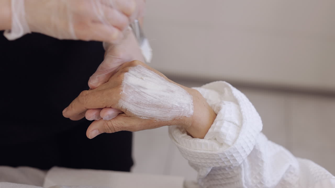 Beautician applying moisturizing medical peeling cream on senior elderly woman hand fingers wrist