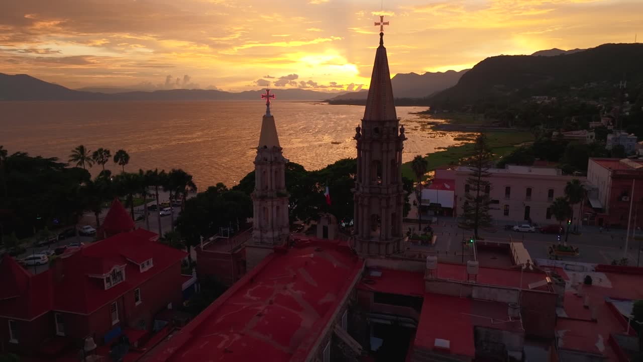 Dolly in flight between the bell towers of Saint Francis Parish in Chapala, Mexico. Chapala Lake at sunset. Orange fiery colors illuminate the lake and mountains