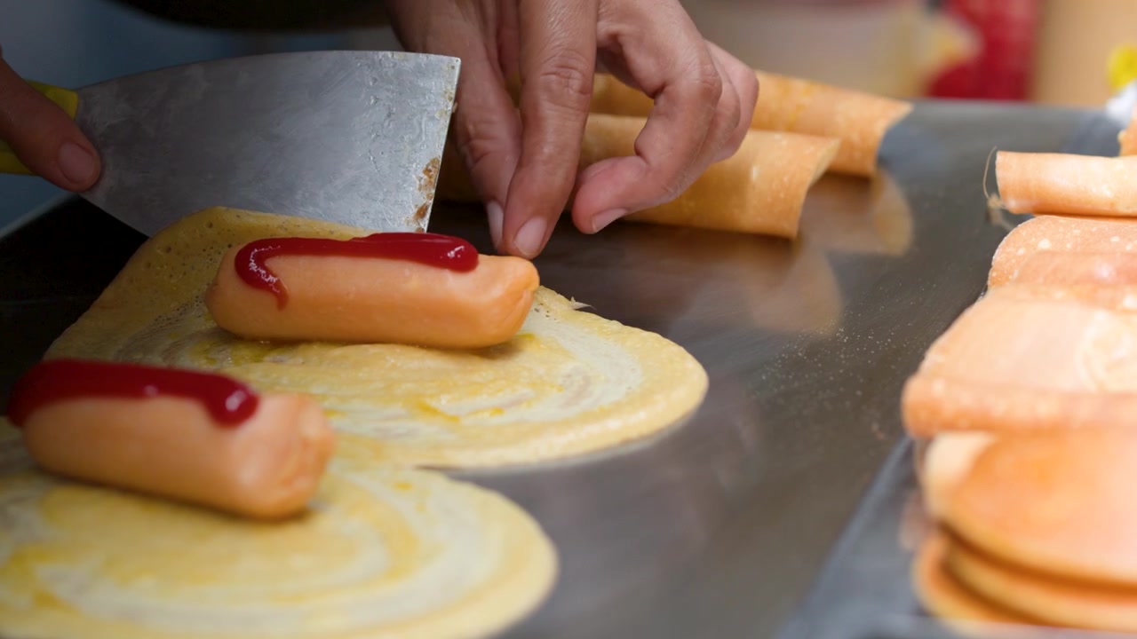 Primo piano di mani che arrotolano frittelle sottili attorno a salsicce con ketchup su una piastra
