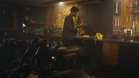 A-brunette-mechanic-guy-in-a-checkered-shirt-starts-his-working-day-puts-on-gloves-and-gets-on-a-motorcycle-in-a-studio-workshop