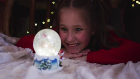 chica caucásica sonriendo y mirando el globo de nieve mientras yacía bajo la manta fuerte durante la navidad en
