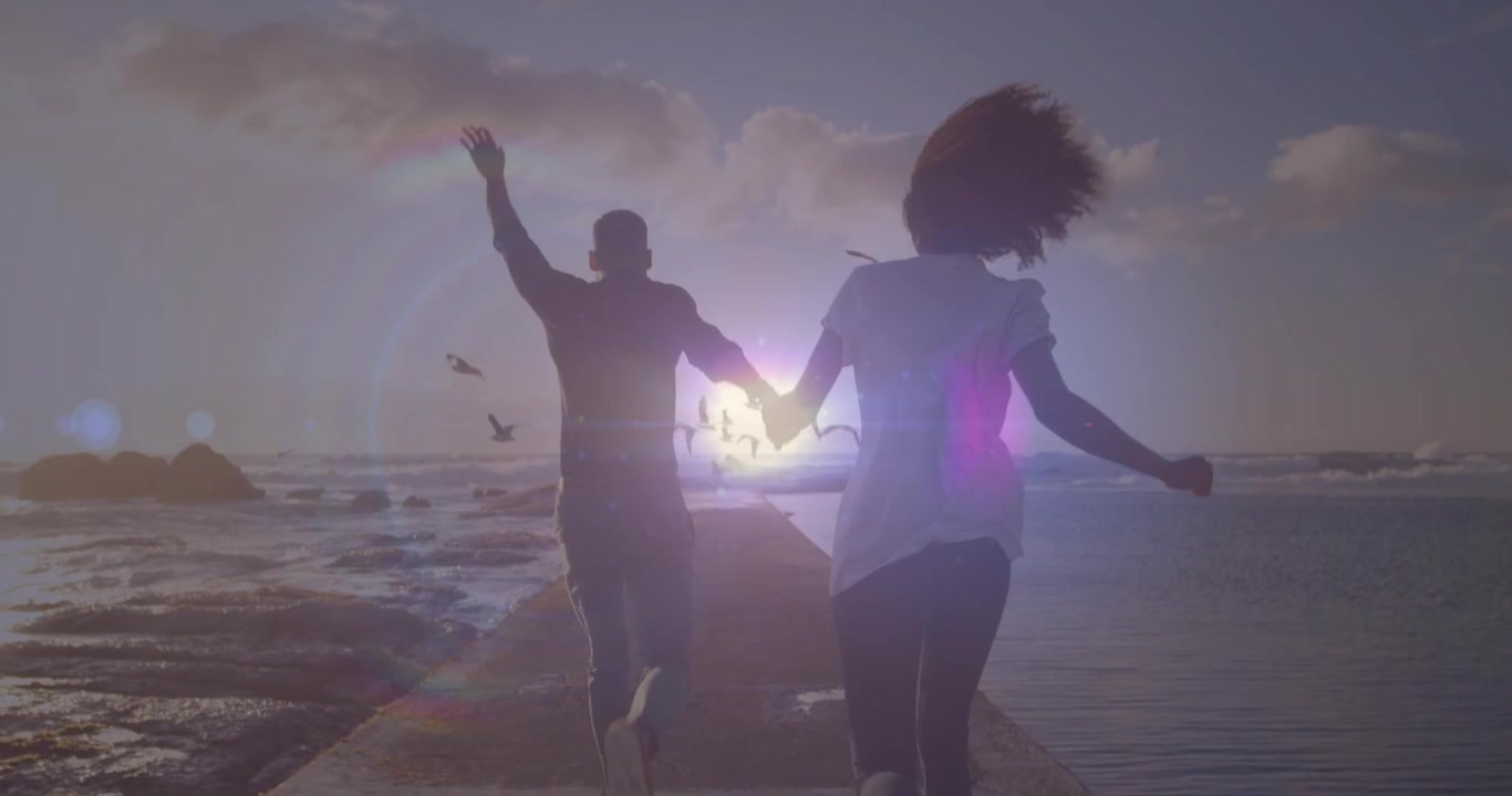Bright spot of light against rear view of african american couple running on the rocks near the sea