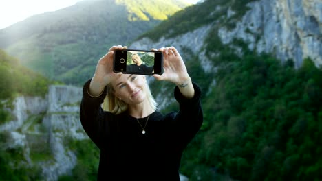 portrait of the beautiful blonde girl taking selfie with her smartphone, holding screen to the camera. in the background scenic green hills and mountains view.