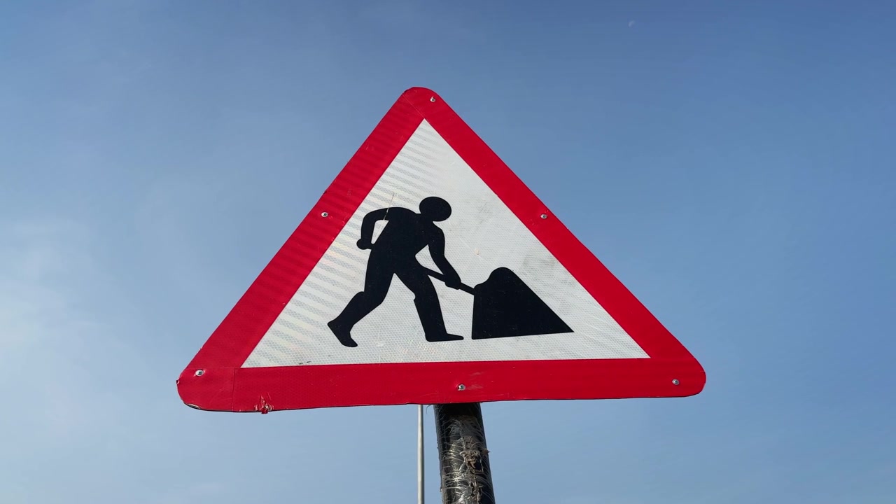 Extreme close up orbiting shot of a triangular road work warning sign of a worker symbol against a clear blue sky The reflective traffic marker emphasizes construction zones and road safety awareness