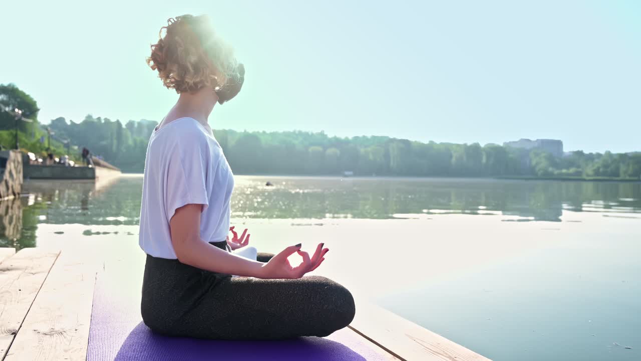 Young curly woman practicing yoga near the lake while wearing black protective mask. Sunny day. Meditating in lotus position on the yoga mat. Corona Virus idea. Chisinau, Moldova