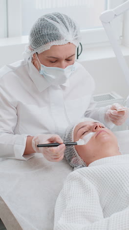 beautician applying moisturizing medical chemical peeling cream mask on young adult woman face