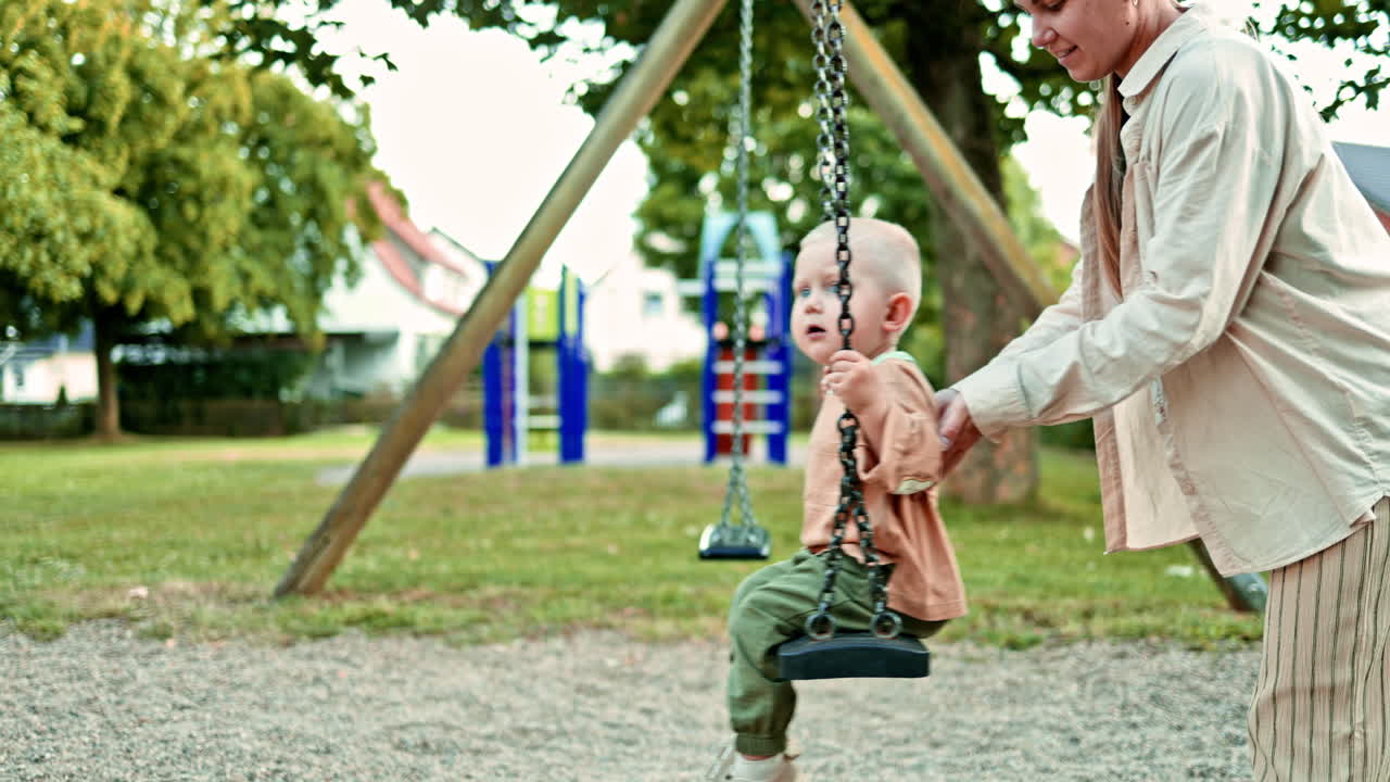 Mamma al parco giochi dondola il suo figlioletto sull'altalena. Il bambino siede sull'altalena e la madre premurosa lo dondola. Mamma e figlio passeggiano all'aria aperta. Concetto di famiglia felice. Sviluppo infantile