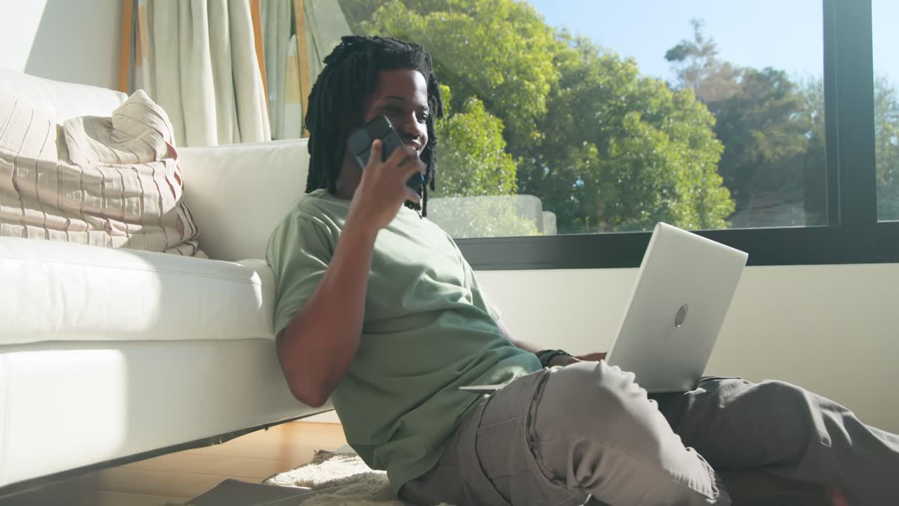 African man sitting on rug by sofa in green tee, typing silver laptop, answering incoming phone