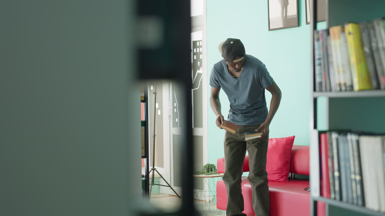 uomo seduto su un divano rosso nel salotto di una biblioteca, gambe incrociate e libro aperto, concentrato, atmosfera tranquilla e studiosa, parete color menta, scaffali, luce dalla finestra, berretto casual, postura rilassata, spazio moderno, momento di lettura nel tempo libero