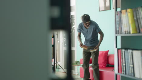 Un hombre se sienta en un sofá rojo en la sala de una biblioteca, cruza la pierna y abre un libro, ambiente de estudio tranquilo y concentrado, pared color menta, estanterías, luz de la ventana, gorra informal, postura relajada, espacio moderno, momento de lectura de relajación