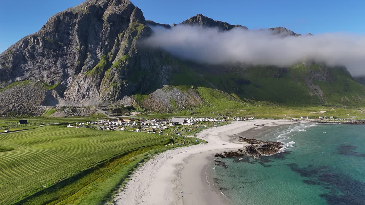 Flakstad strand in Lofoten, dronebeeld, berg bedekt met witte wolken, wit zandstrand en volle camping.