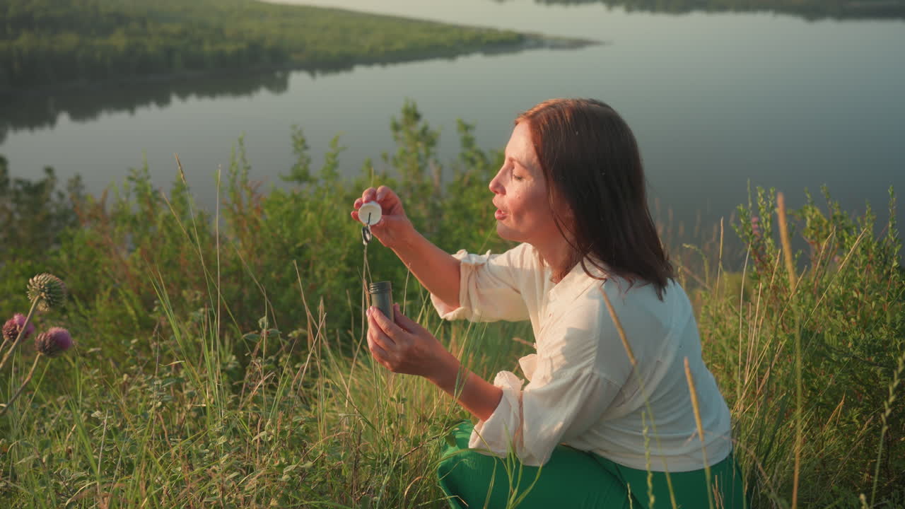 Sunset lit woman in green skirt squatting on grassy river bank blowing shimmering soap bubbles that drift above blurred river valley and wildflowers under warm golden light