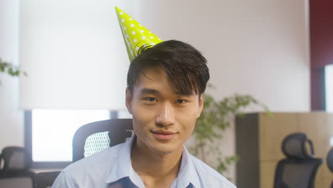 portrait of a smiling man wearing a party hat and looking at camera while confetti falling at the office