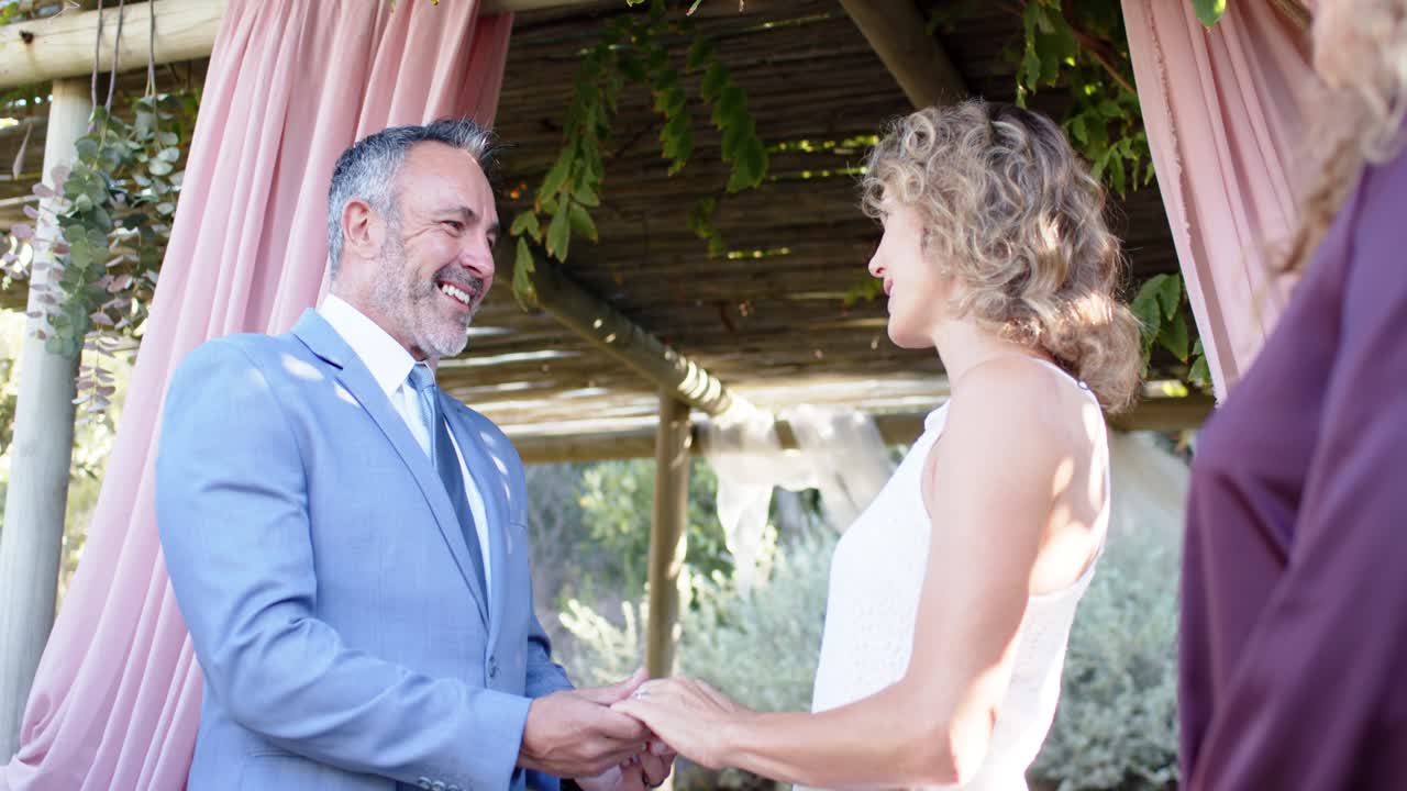 Couple holding hands pink-drape pergola groom speaking vows gowned bride listening Indian officiant
