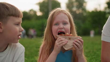 zoom in of energetic girl with playmate in park holding snacks while playfully biting with few teeth, smiling and enjoying outdoor picnic moment on green grass