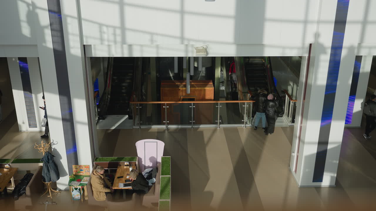 Spacious shopping mall interior featuring people walking and using escalators, modern glass railings, decorative cylindrical ceiling lights, and bright natural daylight casting soft shadows on walls