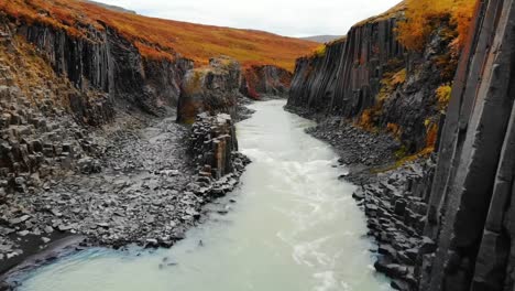 A-4K-drone-captures-cinematic-aerial-footage-of-a-river-winding-through-a-landscape-of-striking-rock-formations,-accented-by-vibrant-orange-hued-grass