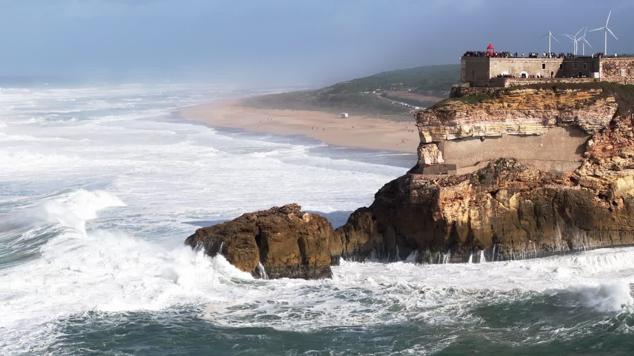 Enormi frangenti atlantici si infrangono sulla costa rocciosa di Nazaré, Portogallo. Drone aereo vicino al faro di Farol da Nazaré e turbine