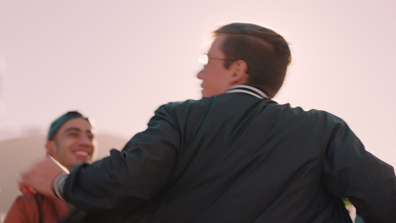 다민족 친구 그룹이 어울려서 젊은 백인 남자가 춤을 추고 해가 지는 날 옥상 파티를 즐기고 술을 마시고 주말 축하를 즐기고 있습니다.