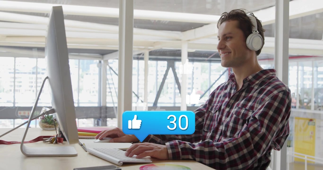 Uomo che digita sulla tastiera in uno studio di design facendo salire "mi piace" blu fluttuanti monitorando l'engagement