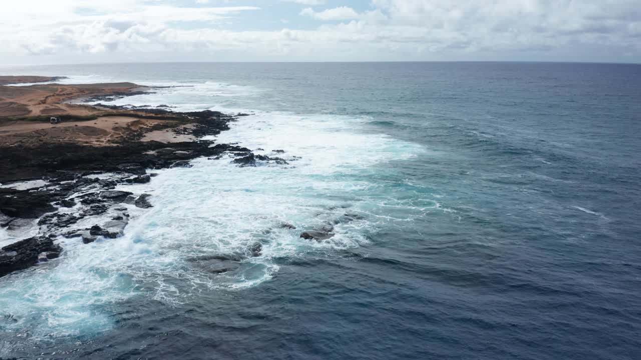 Remote Hawaiian coastline with dramatic black lava rock formations and foamy blue waves crashing ashore beneath a vibrant sky. Aerial view highlights contrast between land and sea