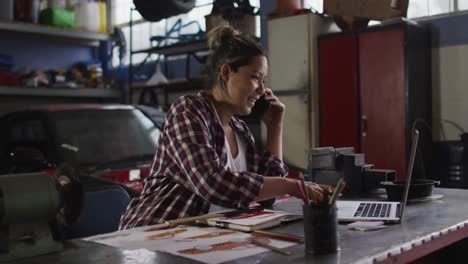 Mecánica-Femenina-Hablando-Por-Teléfono-Inteligente-Y-Usando-Una-Computadora-Portátil-En-Una-Estación-De-Servicio-De-Automóviles