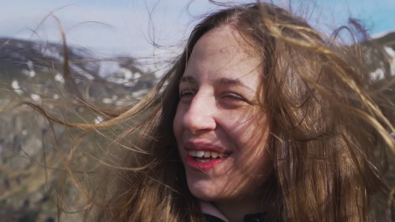 A joyful young woman stands at Botev Peak in Stara Planina, Bulgaria. Captured in slow motion, her hair blows in the wind as she smiles in the vast mountain setting