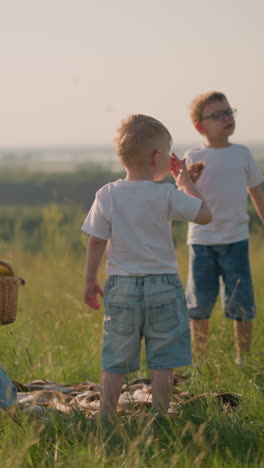 a mother in a blue dress bends down ,takes a basket of fruit while her two young sons, both in white tops, stand nearby in a grassy field