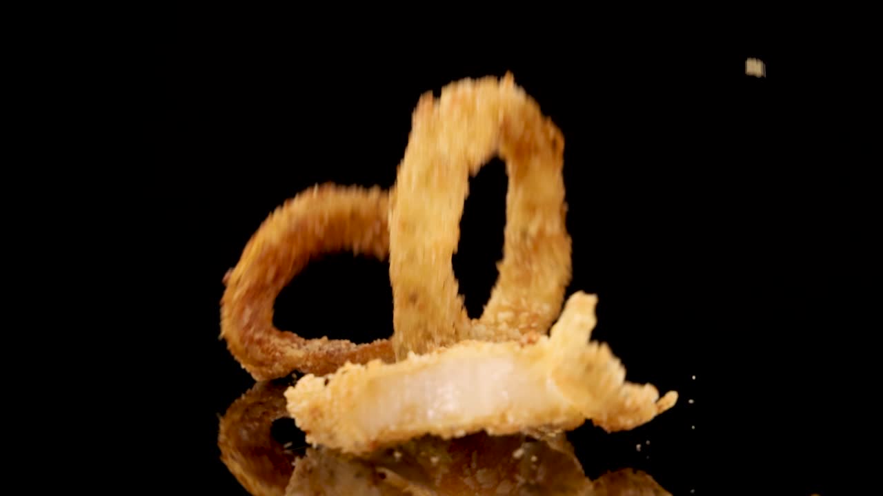 Golden breaded onion rings fall and settle on a reflective black background, captured in slow motion with dramatic studio lighting and high detail