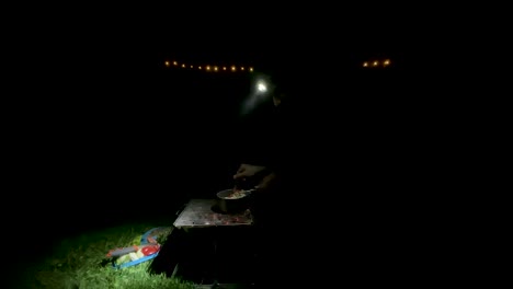 a backpacker cook food on bonfire wood fire barbecue grill at dark night with head light decoration in the wild nature survival