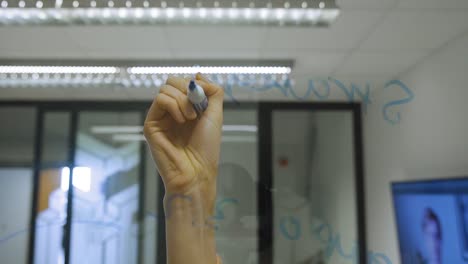 Corporate-professional-young-woman-writes-on-a-glass-window-wall-with-a-marker-during-a-presentation-to-coworkers