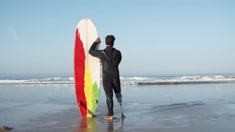 back view of a male surfer in wetsuit leaning on the surfboard and standing in front of the sea looking the waves