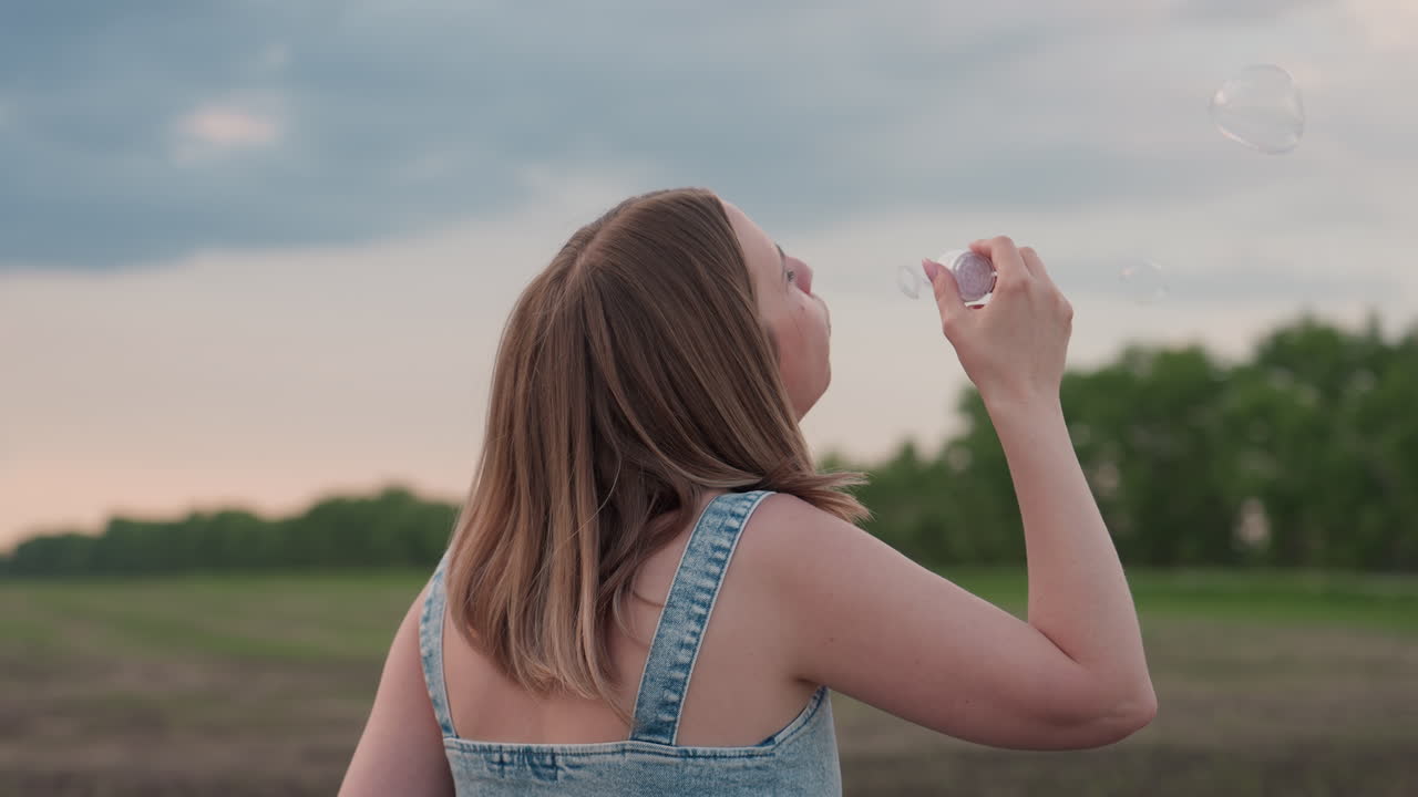 side view of woman walking through farmland blowing bubbles with plastic wand under cloudy sky peaceful summer stroll with playful iridescent spheres drifting in soft breeze over rural dirt road