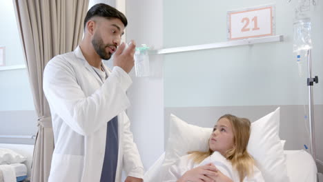 diverse male doctor showing girl patient in hospital bed how to use asthma inhaler, slow motion