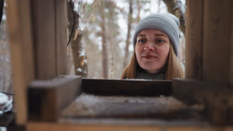 woman wearing gray beanie and winter jacket looks into empty wooden bird feeder in snowy forest, smiling gently, enjoying calm outdoor moment, peaceful winter atmosphere with soft natural light