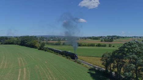 Eine-Drohnenansicht-Eines-Antiken-Dampfpersonenzuges,-Der-In-Zeitlupe-Um-Eine-Kurve-Kommt-Und-An-Einem-Sonnigen-Sommertag-Rauch-Und-Dampf-Durch-Fruchtbare-Maisfelder-Fährt-1