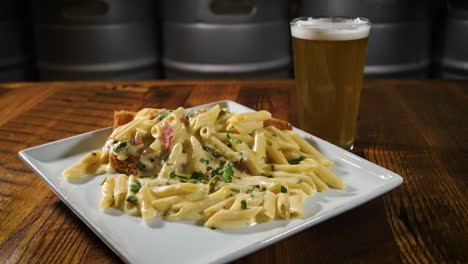 Cremiges-Hühnchen-Alfredo-Mit-Penne-Nudeln-Und-Gebratener-Hähnchenbrust,-Gepaart-Mit-Einem-Pint-Bier-In-Einer-Brauerei,-Slider-4k