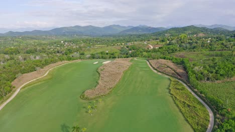 Toma-Aérea-Hacia-Atrás-De-Un-Gran-Club-De-Golf-Con-Campos-De-Hierba-Rodeados-De-Montañas-En-La-Isla-De-República-Dominicana