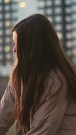 A girl in a peach jacket and black trousers is seen skating towards a bench in an urban park. After sitting down, she rests her hand on the bench, with her face turned slightly away from the camera