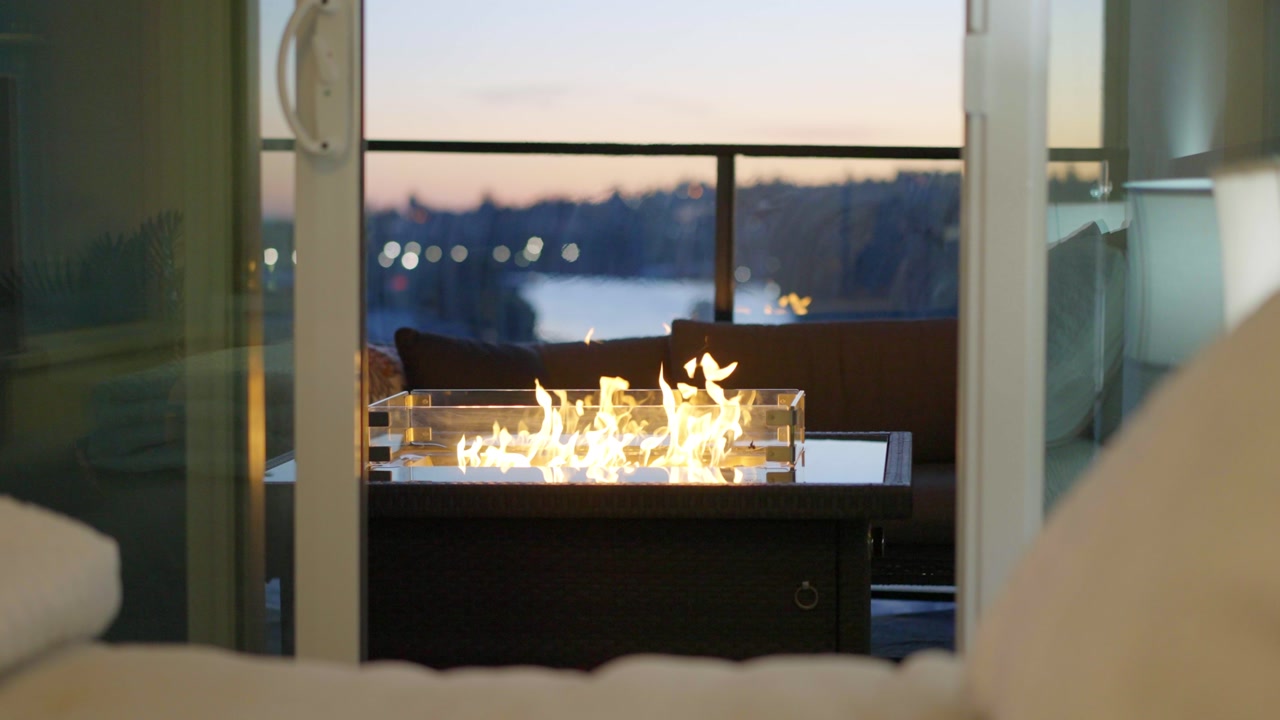 Cozy balcony scene featuring a modern fire table at dusk, viewed from indoors with soft lighting and a peaceful city backdrop.