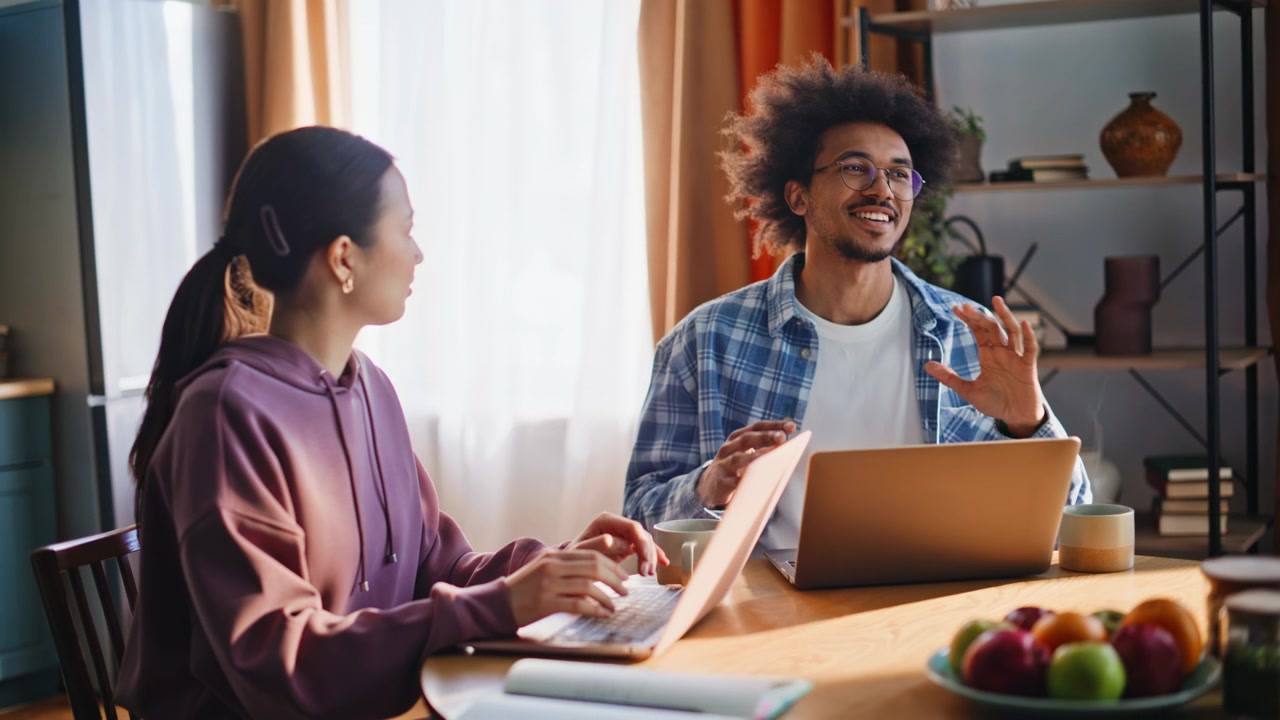 Diverse couple working laptops home kitchen. Man chatting woman typing morning.